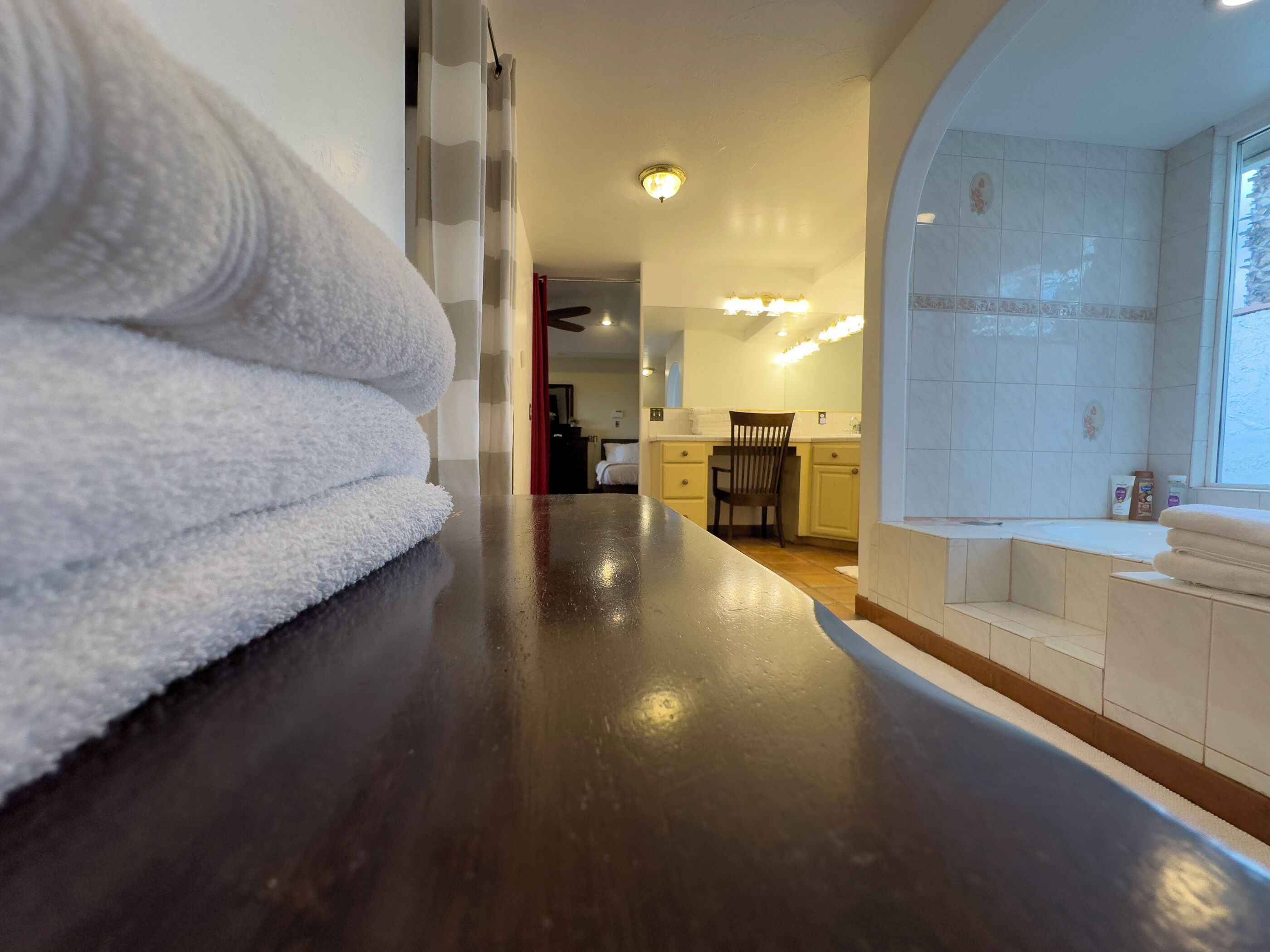 Un pasillo de hotel con suelo de madera, una pila de toallas blancas, una zona de cocina con lavabo y armarios, y un baño con espejo y toallero.