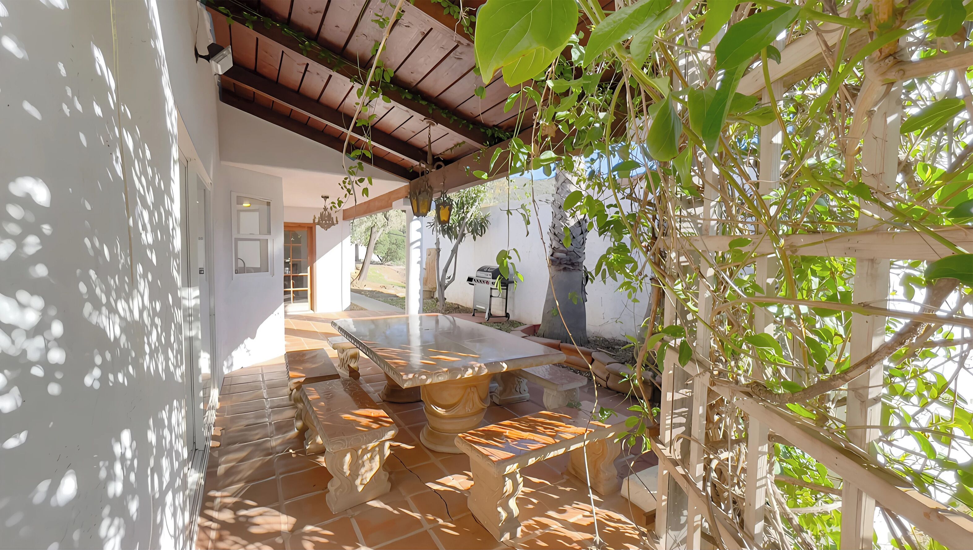 Un patio con mesa y bancos, rodeado de plantas y una pérgola.