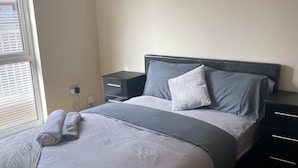 Una cama bien hecha con un edredón gris y blanco, dos almohadas y toallas enrolladas al lado. Hay una mesita de noche con cajones y un enchufe en la pared.