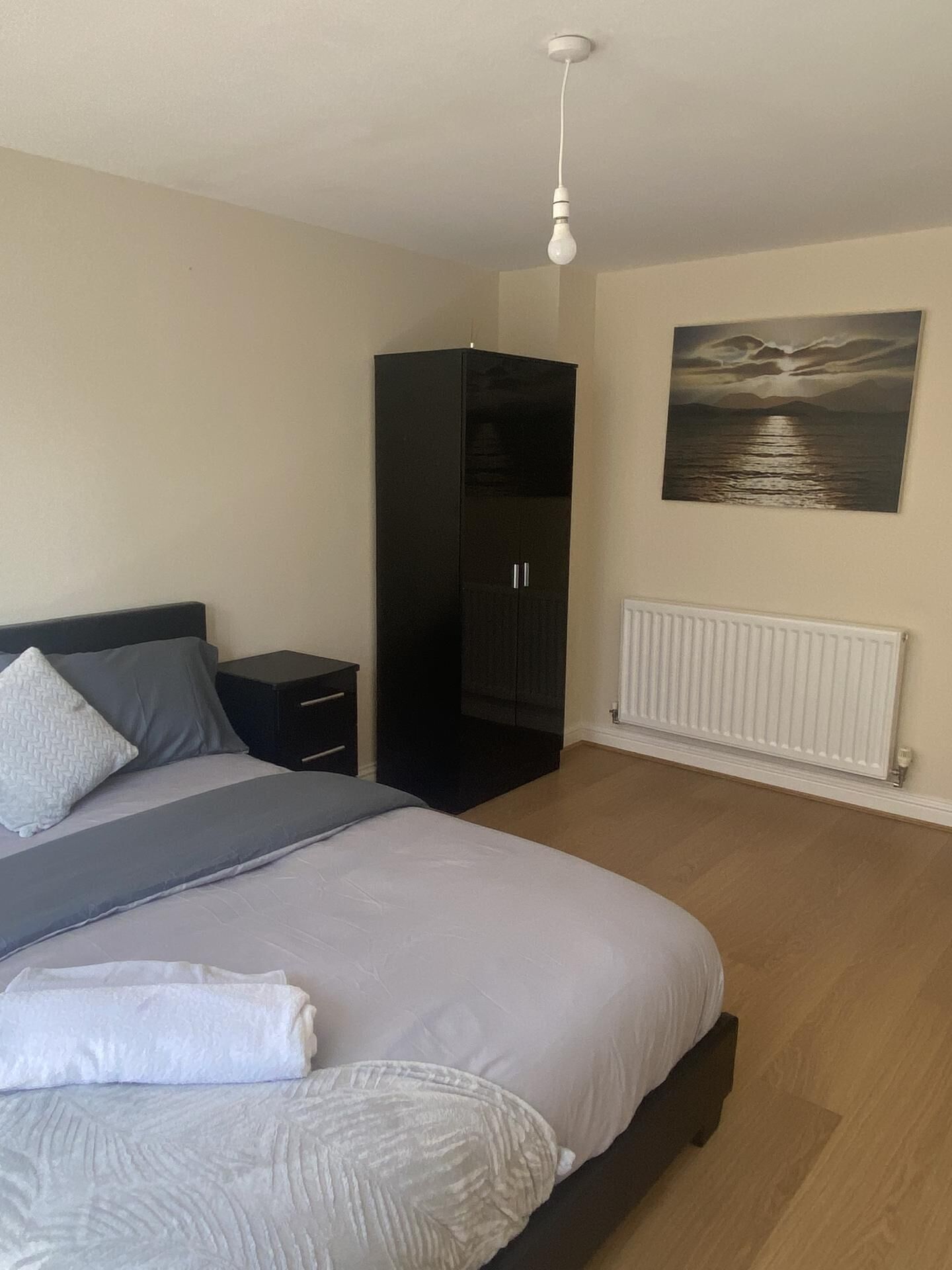 Ein Schlafzimmer mit Bett, Kleiderschrank, Nachttisch, Heizkörper und einem Bild an der Wand.