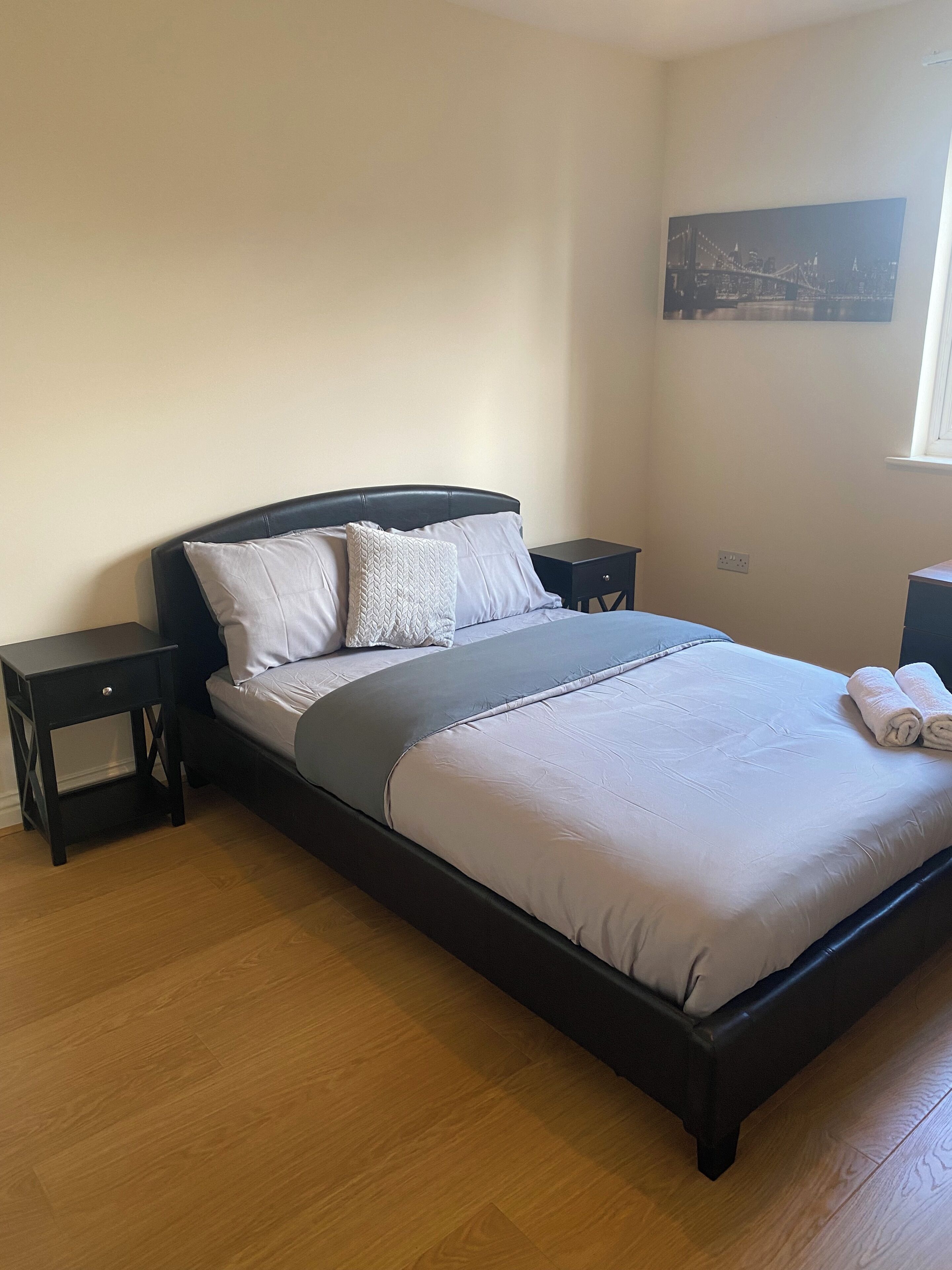 A neatly made bed with a grey and white duvet, two bedside tables, a nightstand, and a wall-mounted picture.