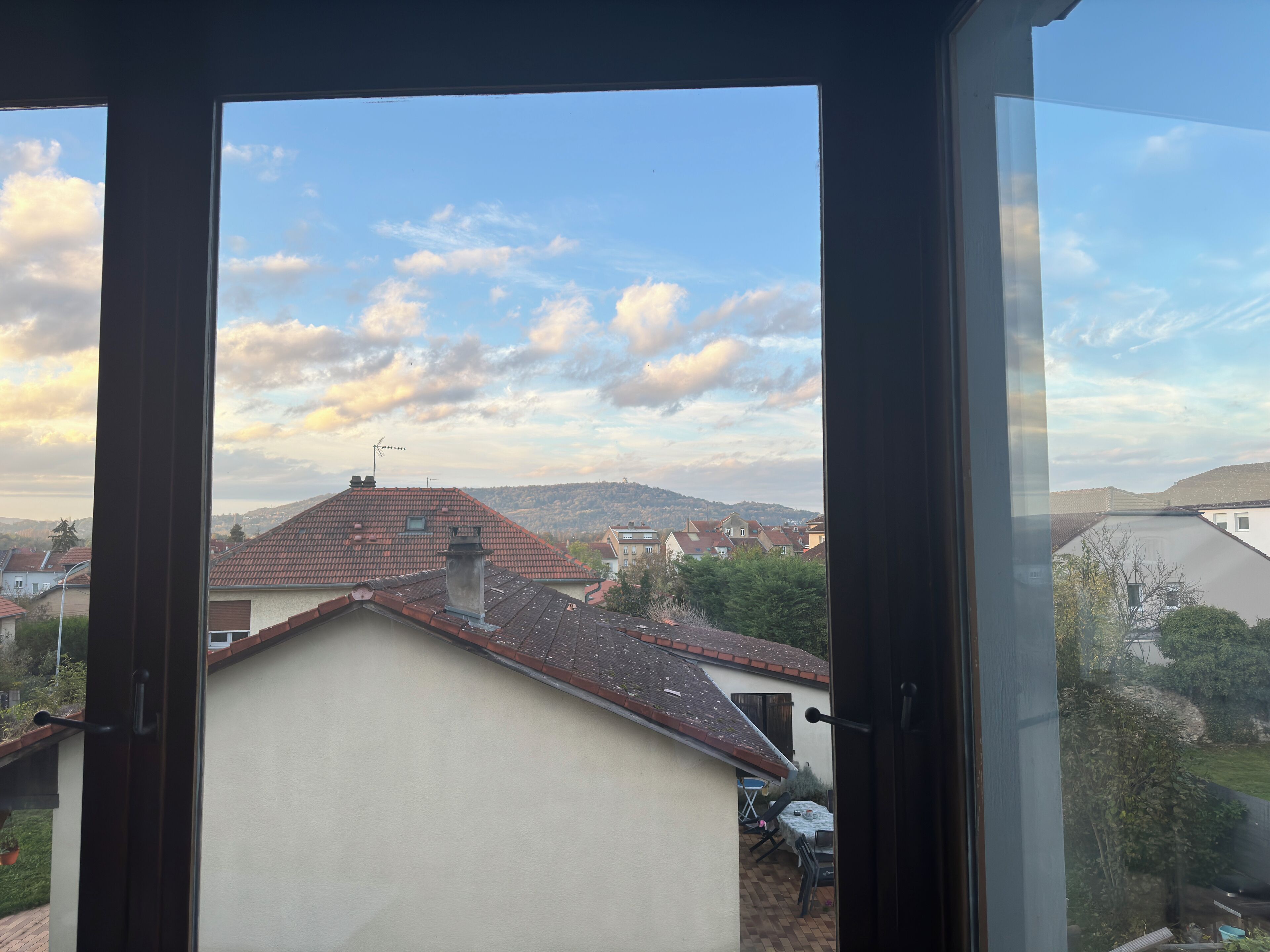 Uitzicht vanuit een raam op een woongebied met huizen, een heldere lucht en een heuvel in de verte.