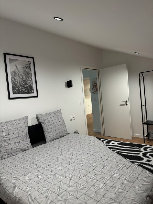 A bedroom with a bed, a framed cityscape photo, a door, and a small black object on the wall.