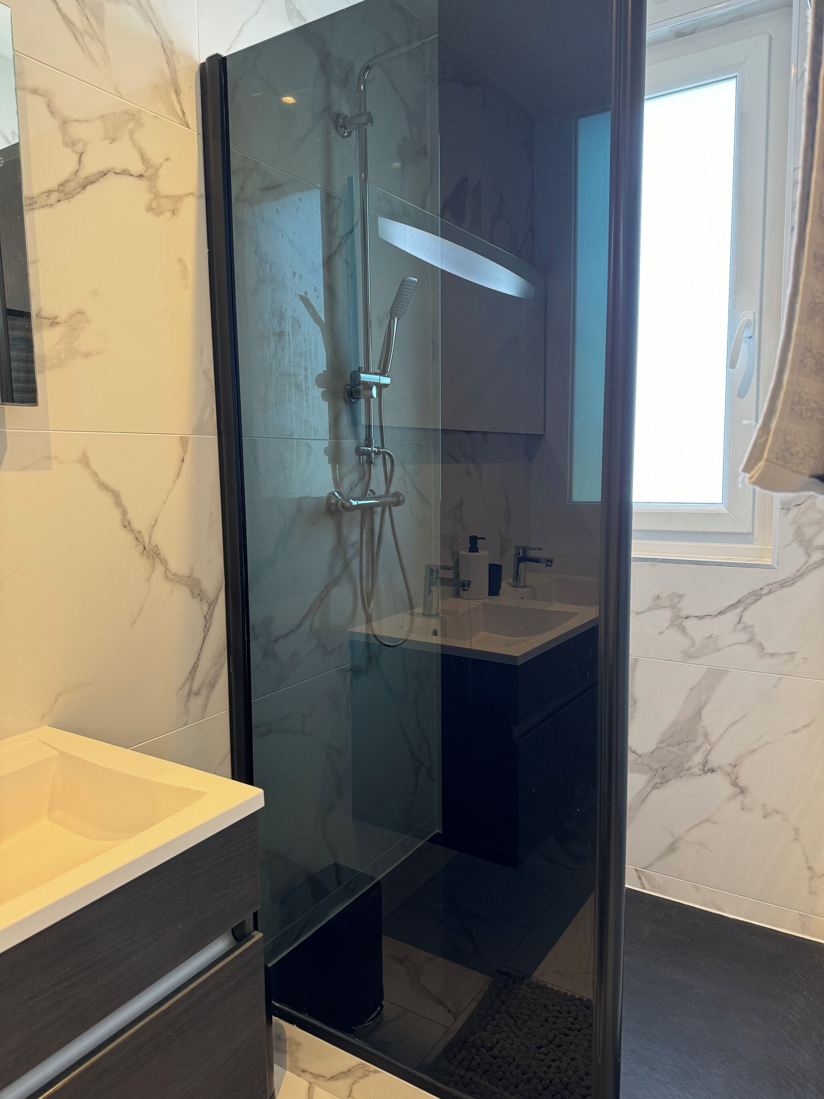 Ein modernes Badezimmer mit einer gläsernen Dusche, Marmorfliesen und einem Waschbecken mit Spiegel.