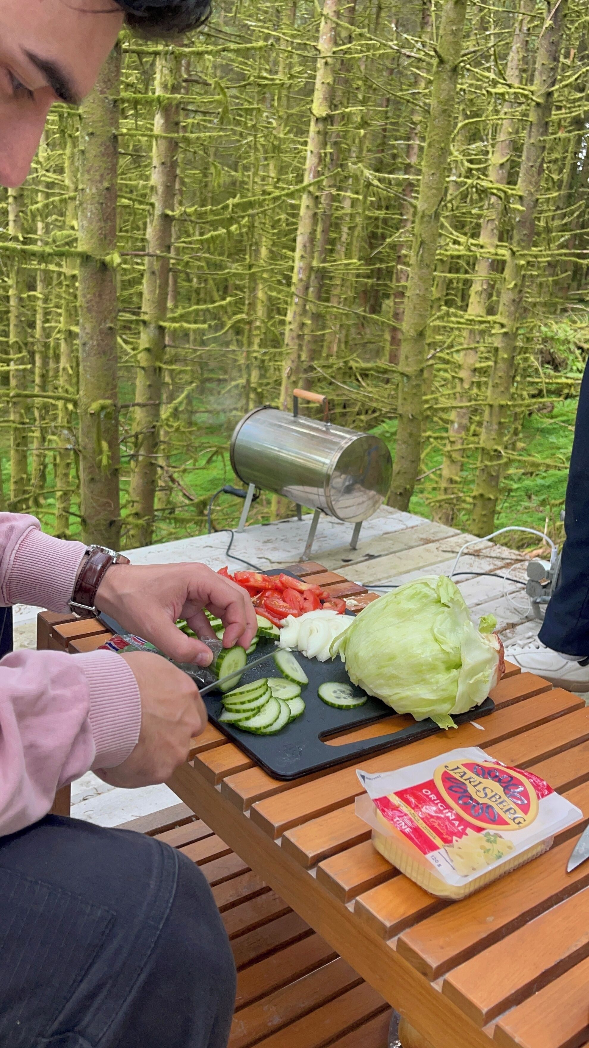 A person slicing vegetables on a wooden table outdoors with a metal grill in the background.