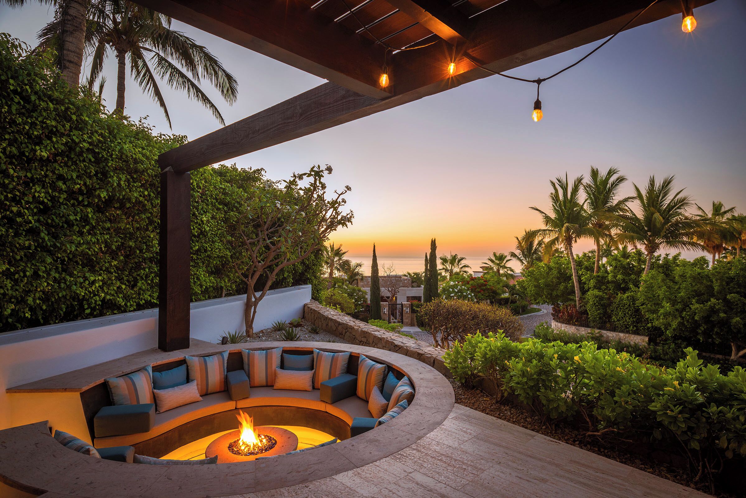 A circular outdoor seating area with a fire pit, surrounded by lush greenery and palm trees, under a wooden pergola with string lights.