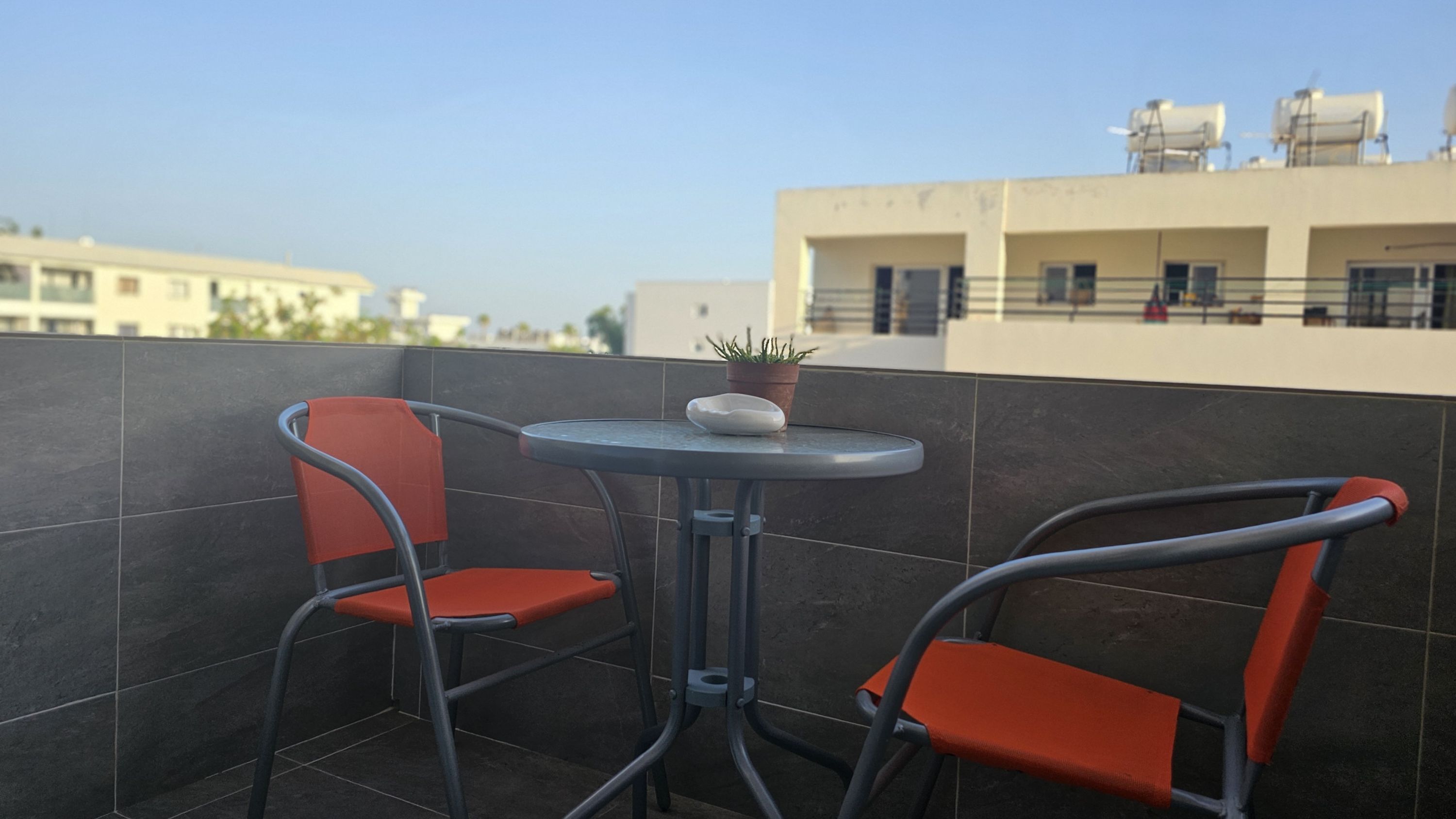 A small outdoor seating area with two chairs and a table, a potted plant, and a view of buildings in the background.