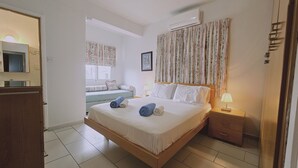 Habitación de hotel con cama, mesita de noche con lámpara, ventana con cortinas, aire acondicionado instalado en la pared y un pequeño rincón de descanso con un banco y cojines.