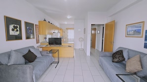 Un salón moderno con un sofá gris, una mesita de cristal y una zona de cocina con armarios de madera y un televisor.