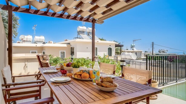 A wooden table set for dining with a view of buildings and a pool in the background. - 114 PRO - Stay Olivia Apartment (Paralimni)