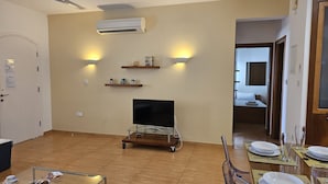 A living room with a flat-screen TV on a cart, two wall-mounted lamps, and an air conditioner. - 114 PRO - Stay Olivia Apartment (Paralimni)