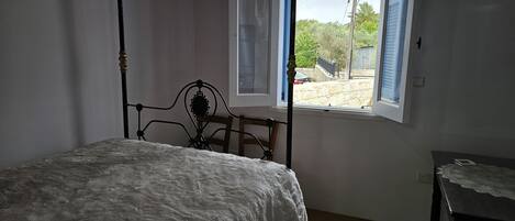 A bedroom with a four-poster bed, a window with a view of the outside, and a small table with a lace tablecloth.