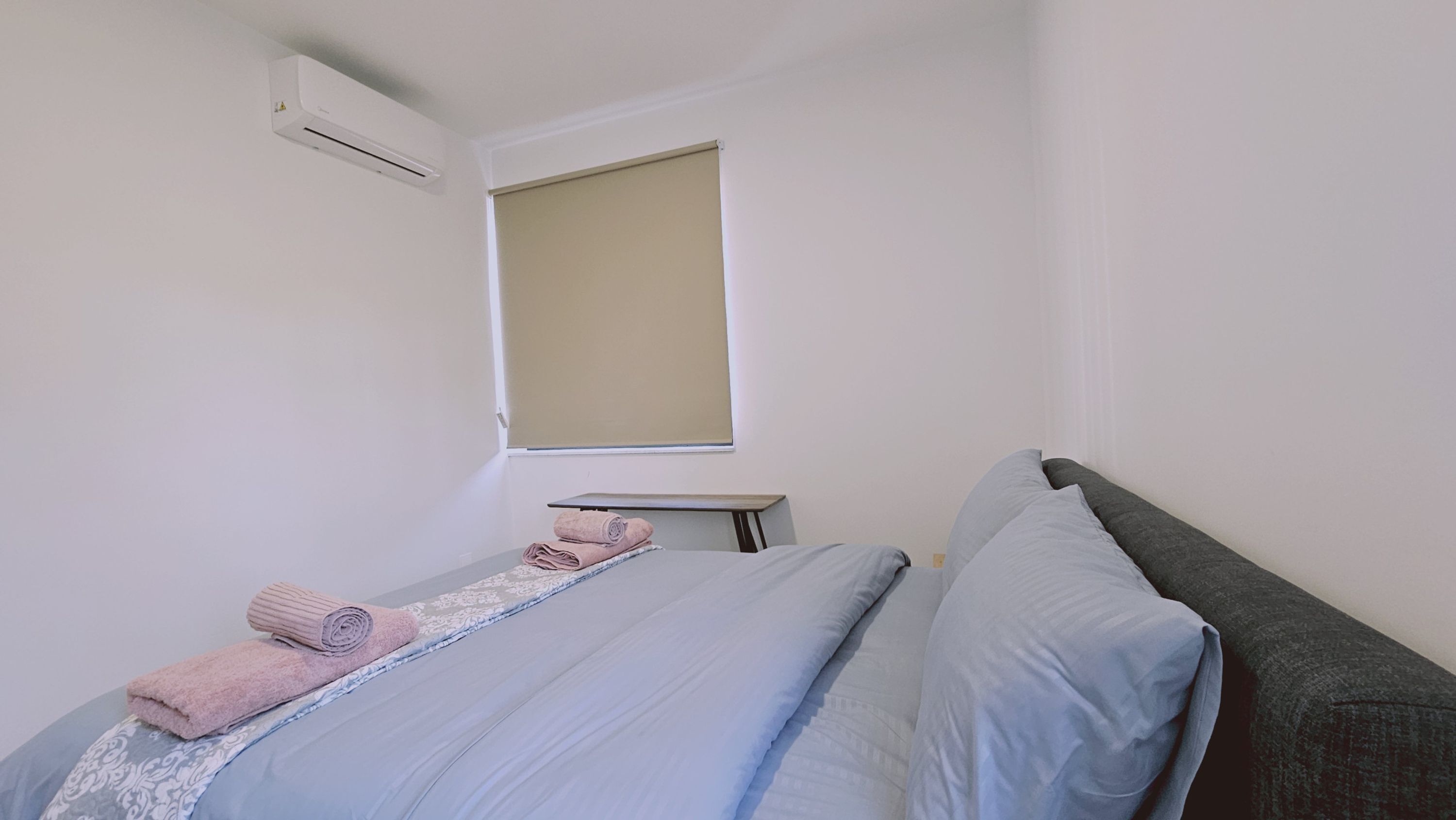 Una camera da letto con un letto, una finestra dotata di un parasole color beige, un comodino con un asciugamano arrotolato e un condizionatore d'aria a parete.