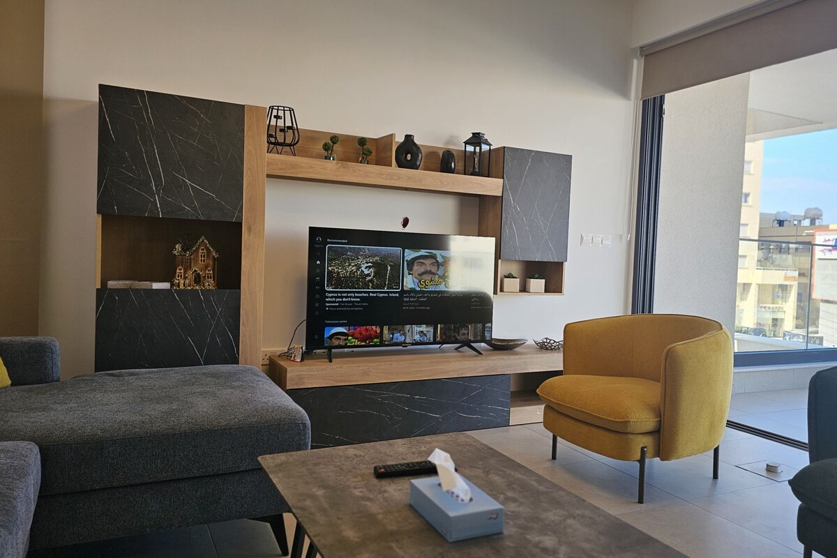 A modern living room with a flat-screen TV, a sofa, a yellow armchair, and a coffee table.