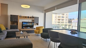 A modern living room with a flat-screen TV, comfortable seating, and a large window offering a city view. - 132 LCA - Stay Diamond Apartment (Larnaca)