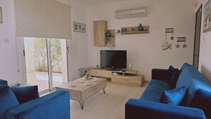 Sala de estar com televisão, sofás azuis, mesa de centro de madeira e uma porta de vidro deslizante que dá para um pátio.