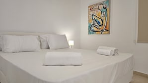 Una cama bien tendida con sábanas y almohadas blancas, una toalla enrollada y un cuadro en la pared.