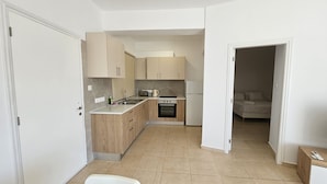 Una cocina compacta con armarios de madera, horno empotrado y refrigerador. Se observa un pequeño comedor al fondo.