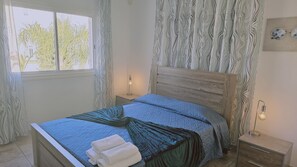A bedroom with a wooden headboard, a bed with blue bedding, a nightstand with a lamp, and a view of trees through the window.