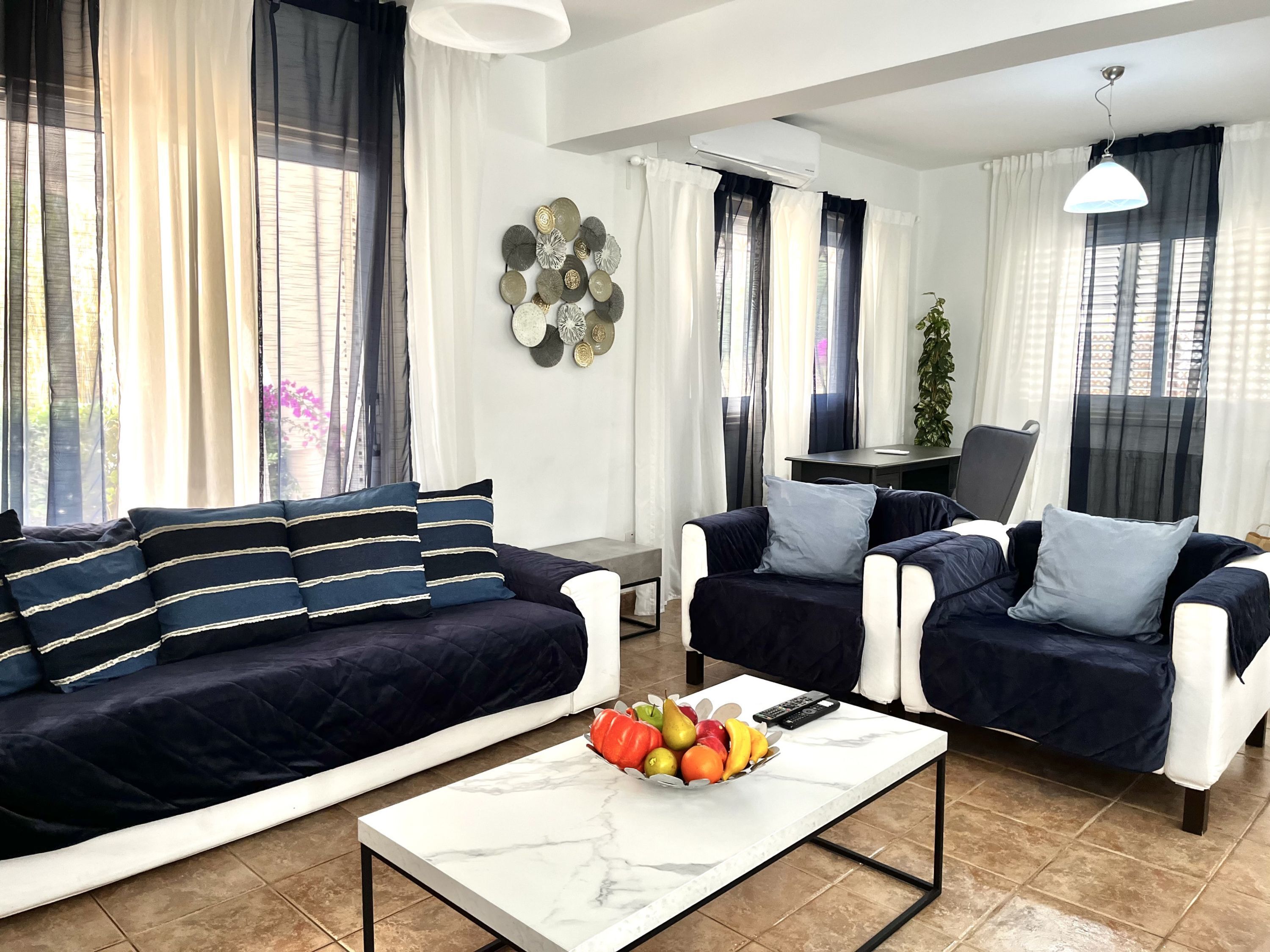 A modern living room with a sofa, two armchairs, a coffee table with fruit, and a desk with a chair.