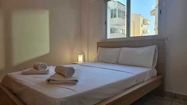 Una cama bien hecha con sábanas blancas y dos toallas enrolladas, una mesita de noche con una lámpara, y una ventana con vistas a edificios.