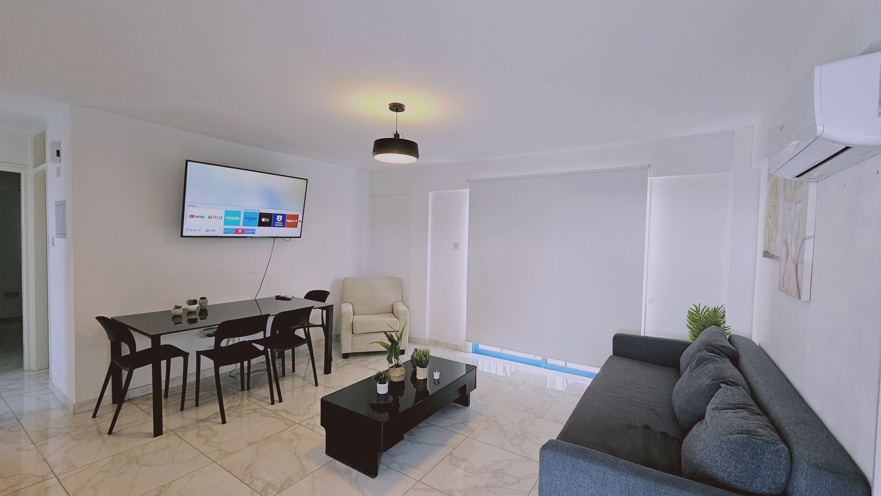 A modern living room with a sofa, coffee table, dining table with chairs, a TV mounted on the wall, and a large window.