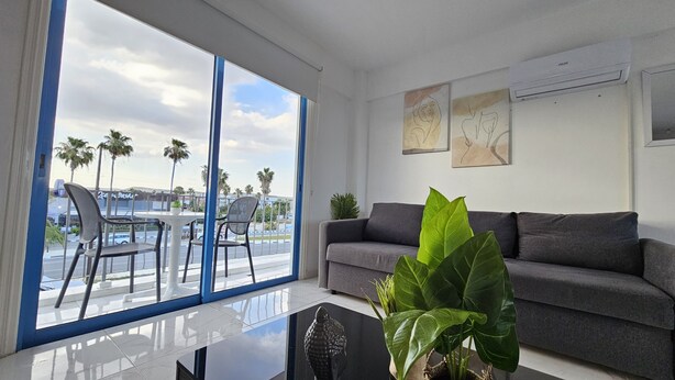 A modern living room with a grey sofa, a glass coffee table, and a view of palm trees and a body of water through large windows. - 140 VOR - Stay Chrisoula Beach Garden (Oroklini)