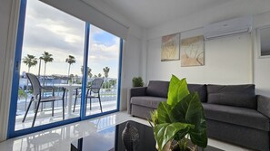 Un salon moderne avec un canapé gris, une table basse en verre et une vue sur des palmiers et une étendue d’eau, le tout visible à travers de grandes fenêtres.
