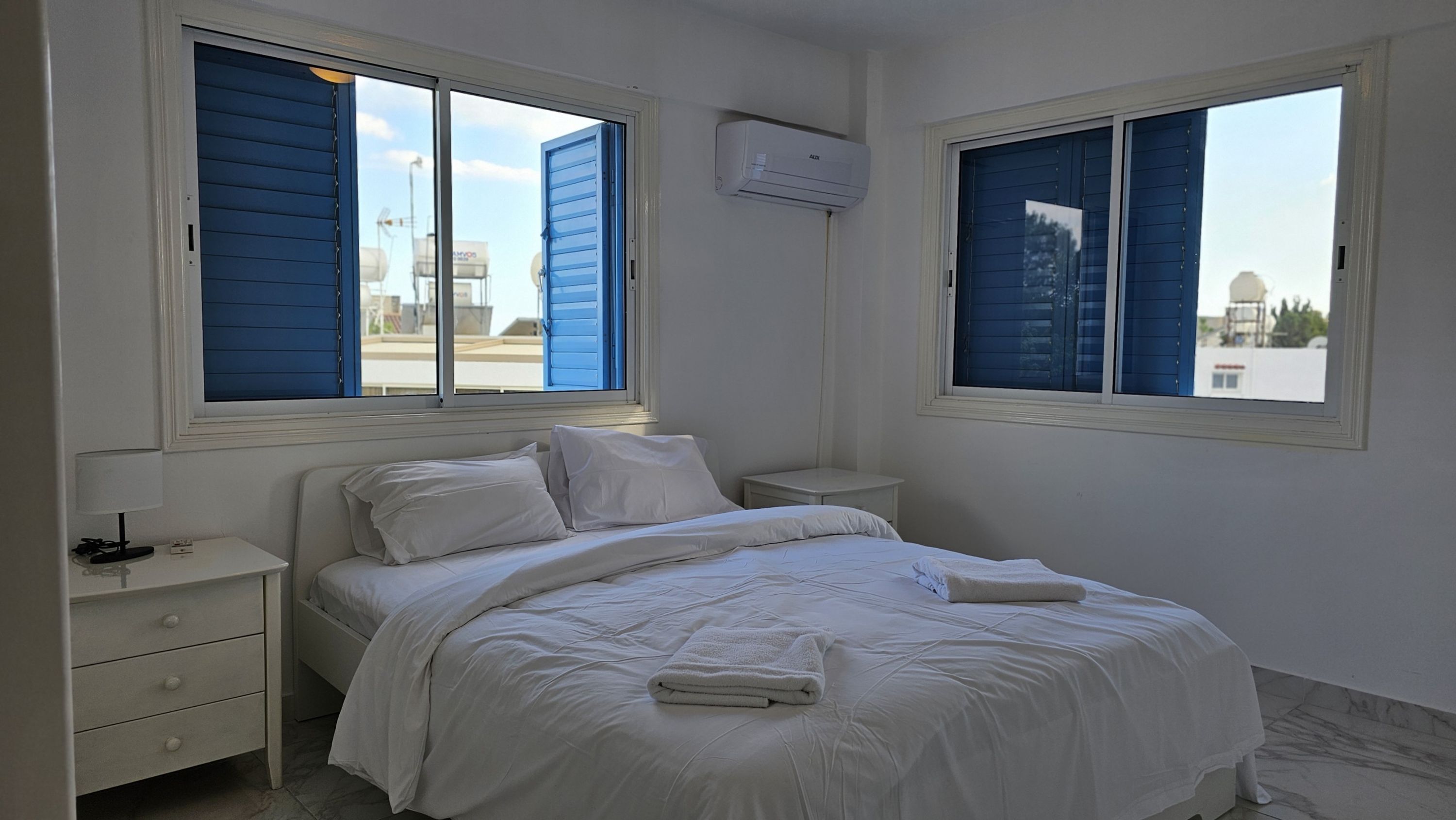 Une chambre à coucher avec un lit, deux tables de chevet, une lampe et deux fenêtres avec des volets bleus.