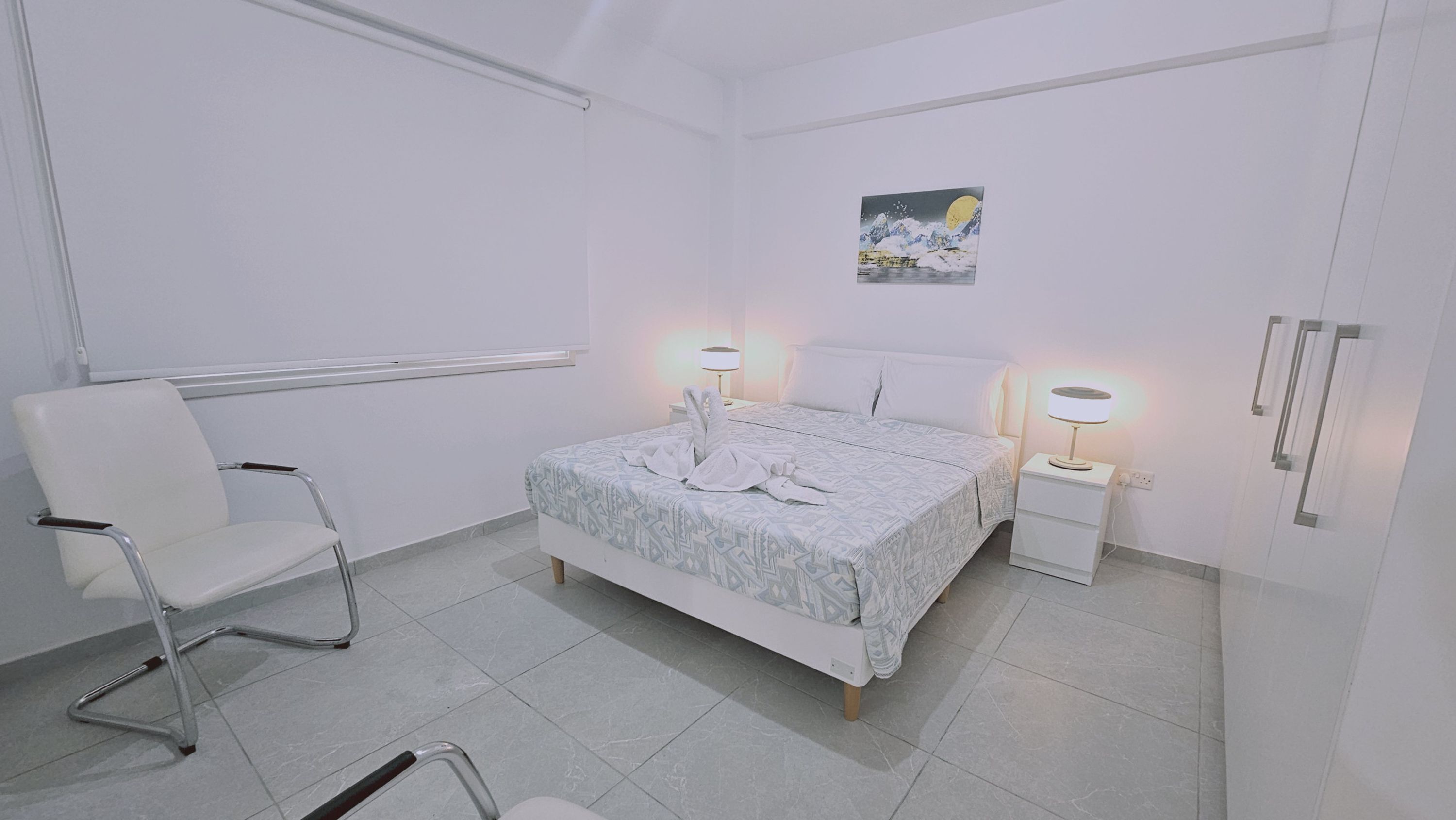 A hotel room with a bed, two bedside tables with lamps, a chair, and a wall-mounted artwork.
