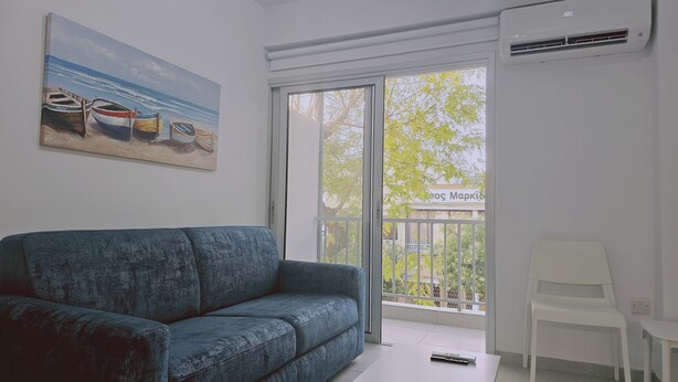 A living room with a blue sofa, a painting of boats, a white chair, and a balcony with a view of trees and a building. - 145 NIC - Stay Historia Apartment (Nicosia)