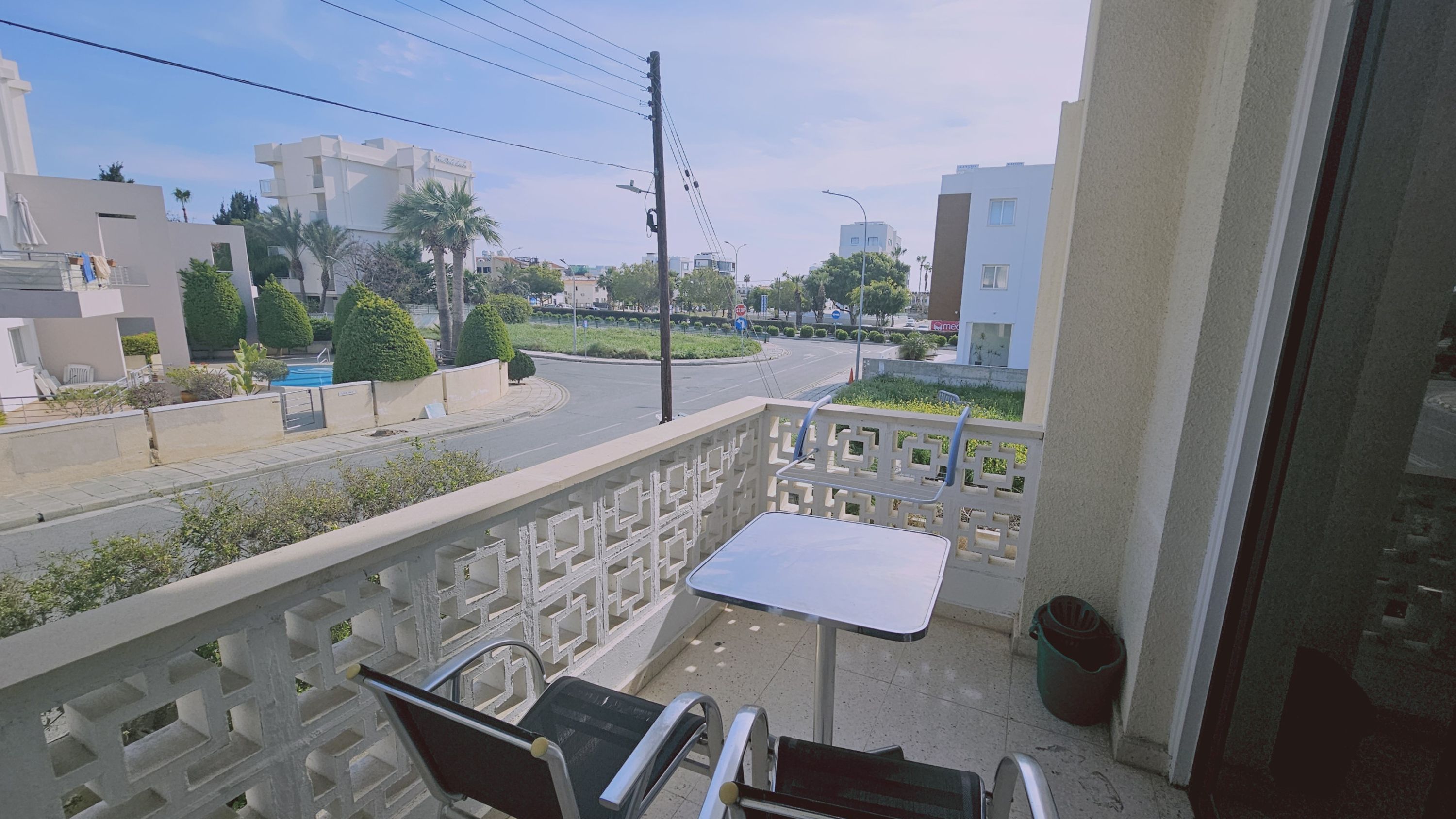 A balcony with a table and chairs, overlooking a road with palm trees and buildings.