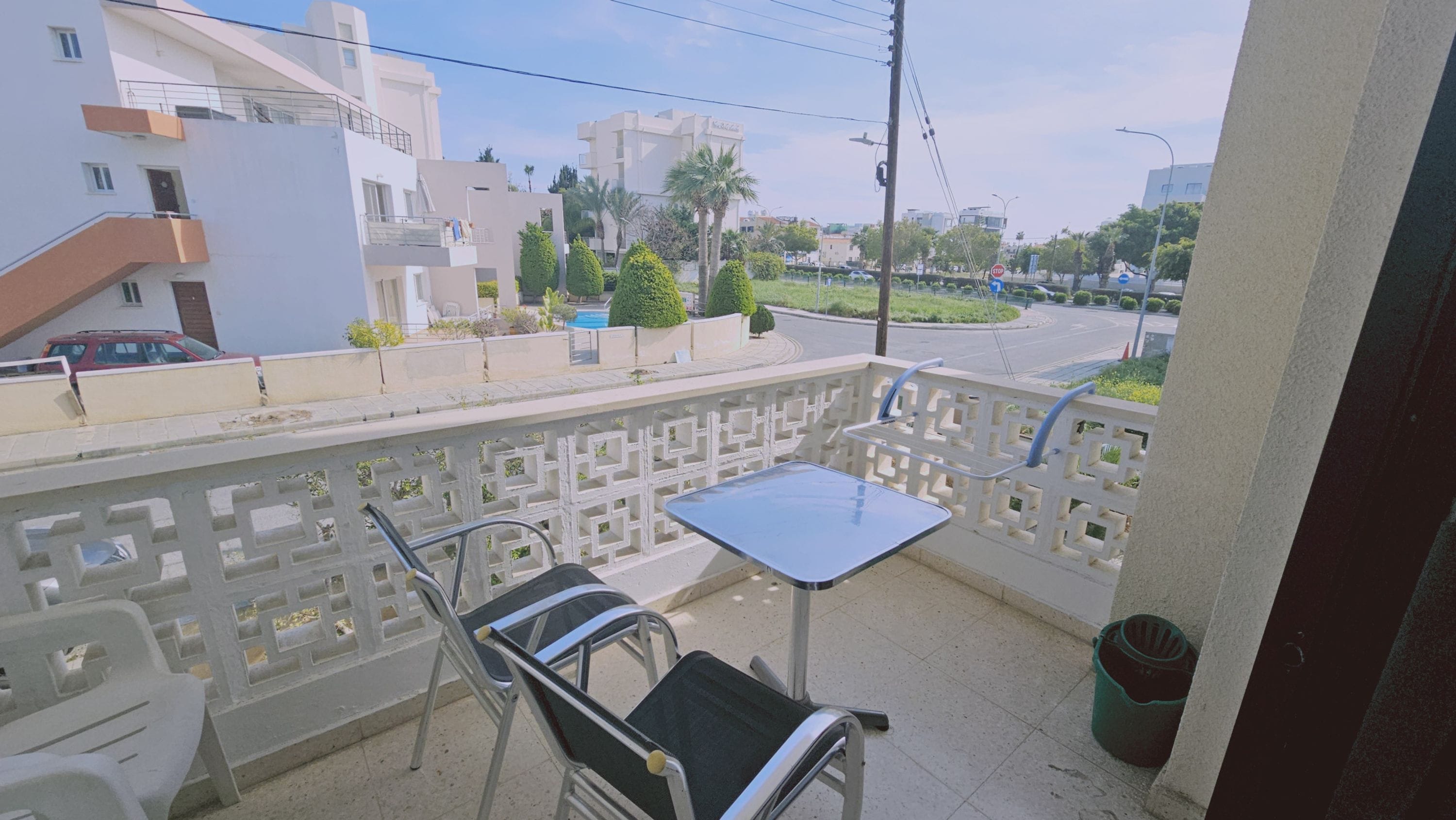 A balcony with a table and chairs, a view of a residential area with buildings and a roundabout, and a stop sign in the distance.