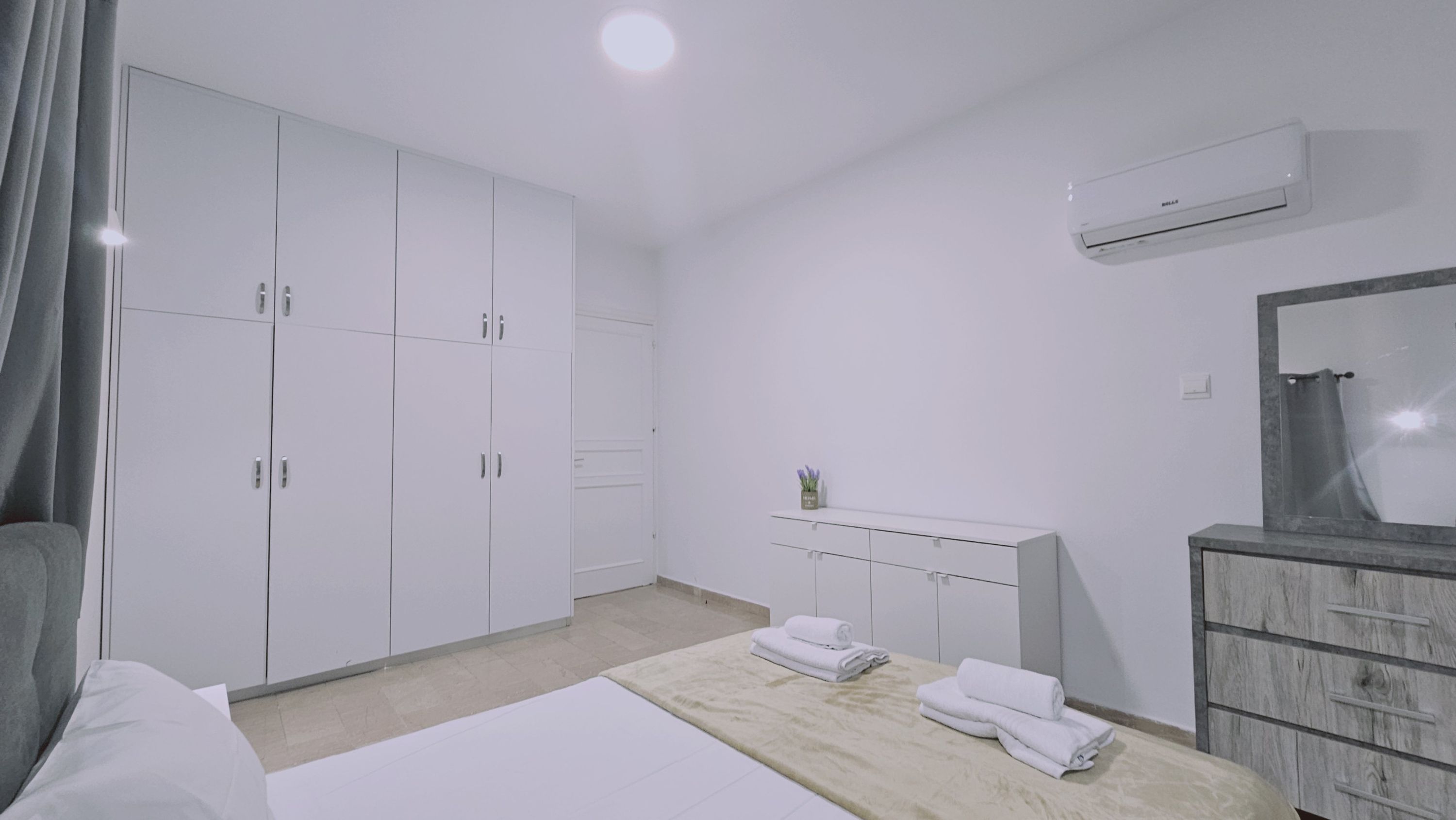 A hotel room with a bed, a wardrobe, a vanity with a mirror, and an air conditioning unit.