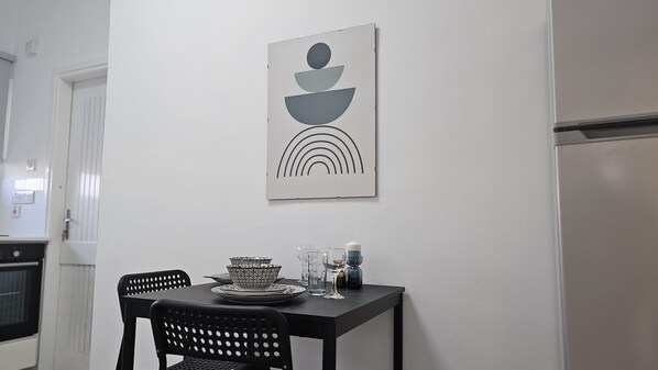 A dining area with a table set for two, a decorative wall piece above the table, and a kitchen area in the background. - 142 NIC 1 - Stay City Life 1 Apartment (Nicosia)