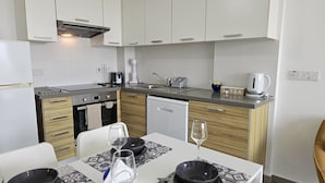 A modern kitchen with a built-in oven, dishwasher, and a coffee maker. There is a dining table with a placemat and wine glasses. - 135 PRO - Stay Arpa Suite (Paralimni)