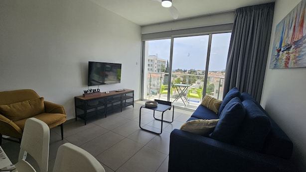 A modern living room with a sofa, armchair, TV, and a balcony with a city view. - 135 PRO - Stay Arpa Suite (Paralimni)