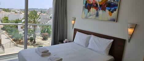 A hotel room with a large bed, two bedside lamps, a painting, and a view of the city.