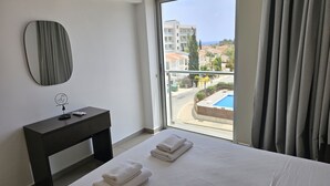 A hotel room with a bed, a bedside table with a remote control, a mirror, and a view of a pool and buildings. - 135 PRO - Stay Arpa Suite (Paralimni)