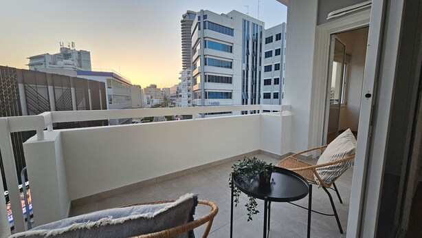 A balcony with wicker chairs and a small table, overlooking a cityscape with modern buildings. - 142 NIC 2 - Stay City Life 2 Apartment (Nicosia)