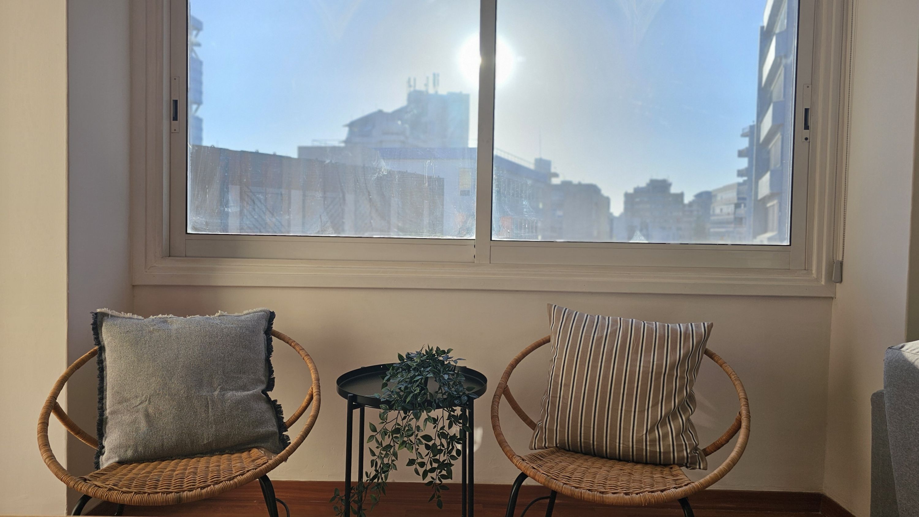 Ein Zimmer mit zwei Korbstühlen, einer mit grauem Kissen und der andere mit gestreiftem Kissen, vor einem Fenster mit Blick auf Gebäude.