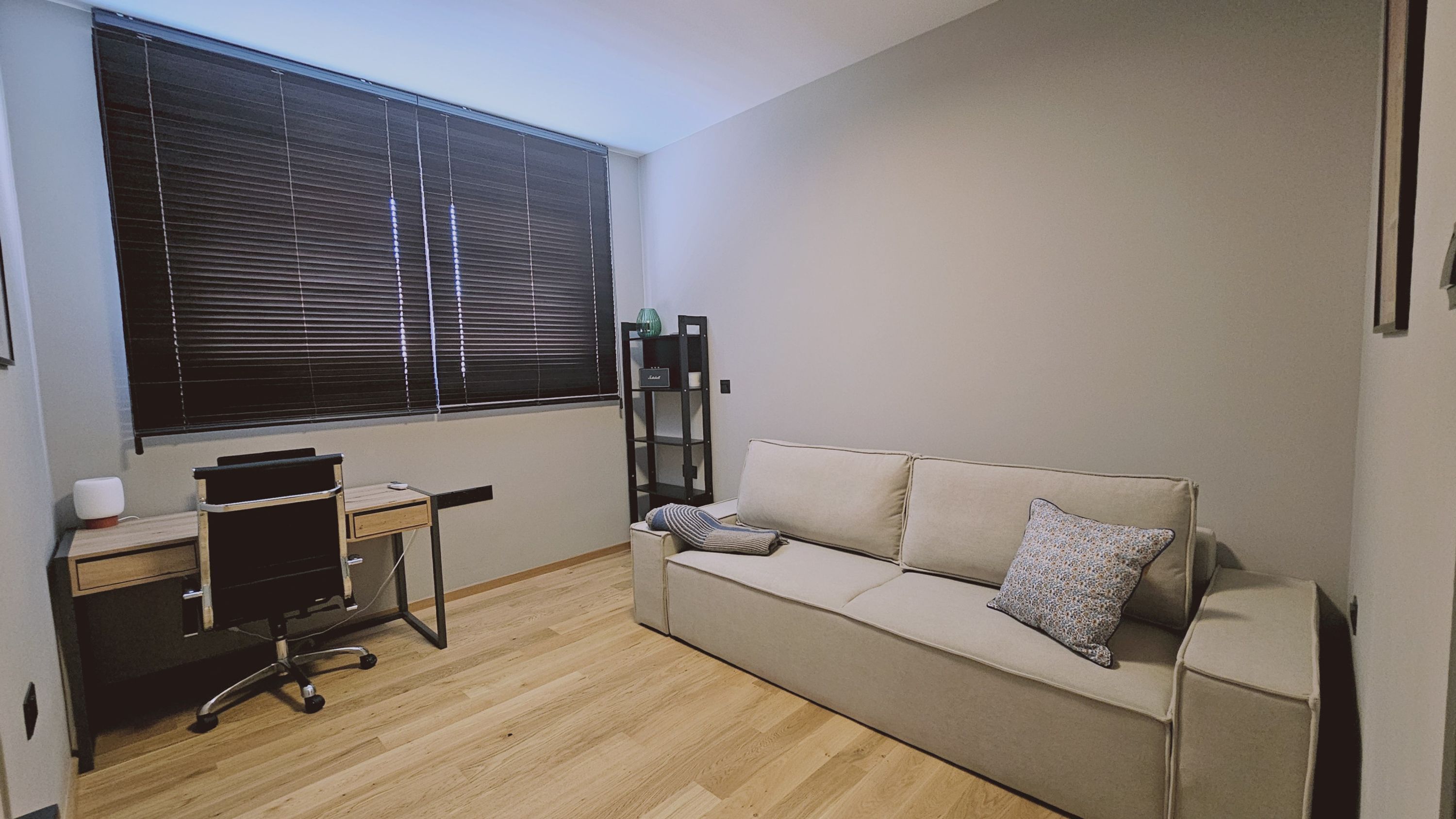 A small, well-lit room with a wooden floor, a beige sofa with a pillow, a desk with a chair, and a window with blinds.
