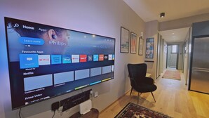 Una sala de estar con un televisor de pantalla plana grande que muestra una pantalla de inicio con iconos de aplicaciones, una silla negra y piso de madera.