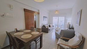 Un salon moderne comprenant un coin repas, un canapé et un fauteuil. Une porte donne accès à une autre pièce, et une fenêtre offre une vue agréable.