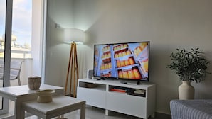 Un salon moderne avec un téléviseur à écran plat posé sur un meuble blanc, un lampadaire, une table à manger avec des chaises, une plante en pot et une vue sur le balcon extérieur.