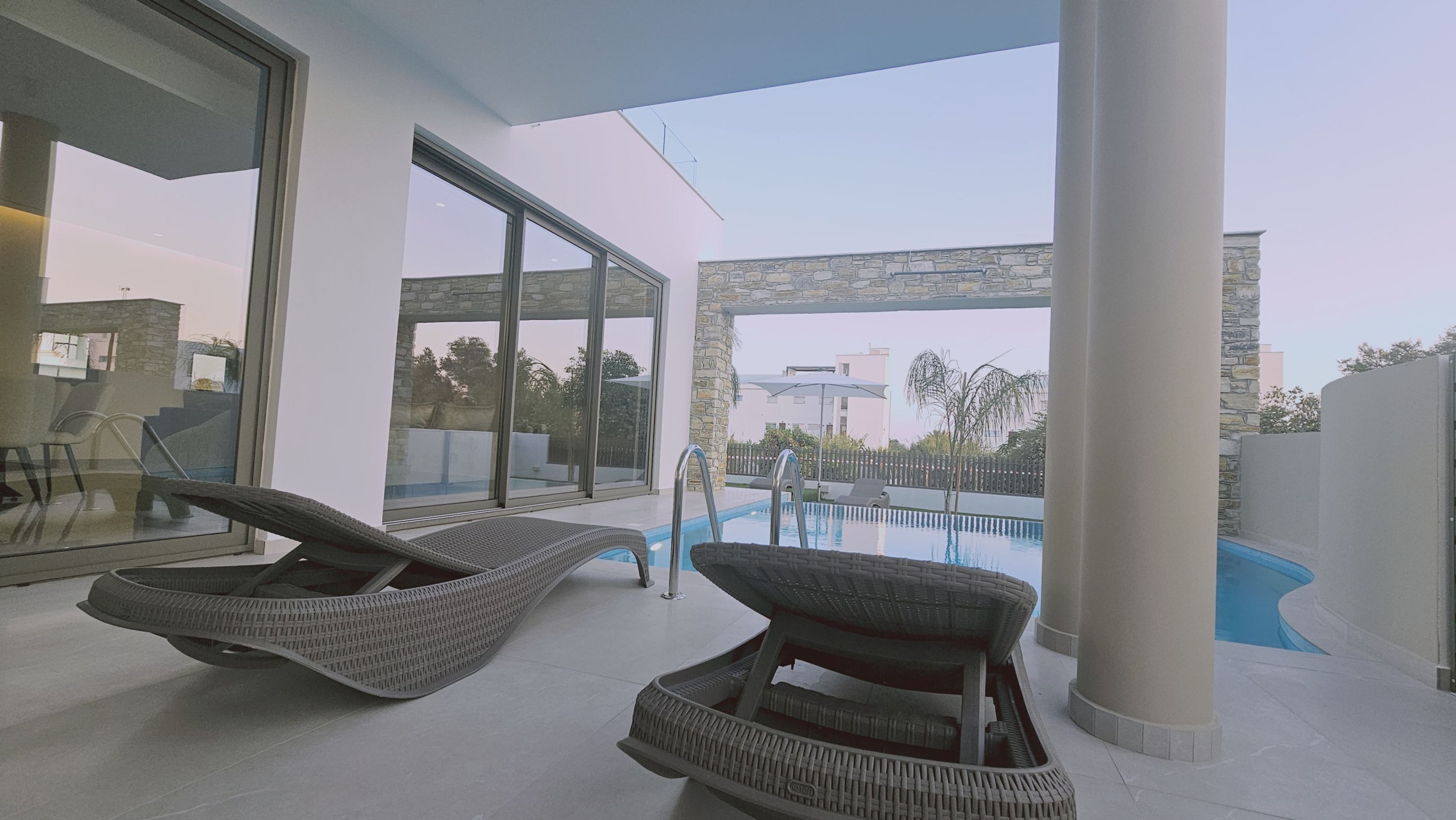 A modern outdoor pool area with two wicker lounge chairs, a clear view of the pool, and a stone wall backdrop.