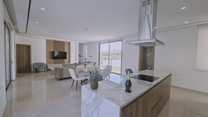 A modern kitchen with a marble island, a dining area with a glass table, and a living room with a TV and wooden panel wall. - 146 4 AN - Stay Oasis Luxury Villa (Ayia Napa)