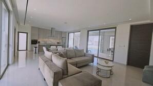 A modern living room with a sectional sofa, a round coffee table, and a view of the kitchen and outdoor area. - 146 4 AN - Stay Oasis Luxury Villa (Ayia Napa)