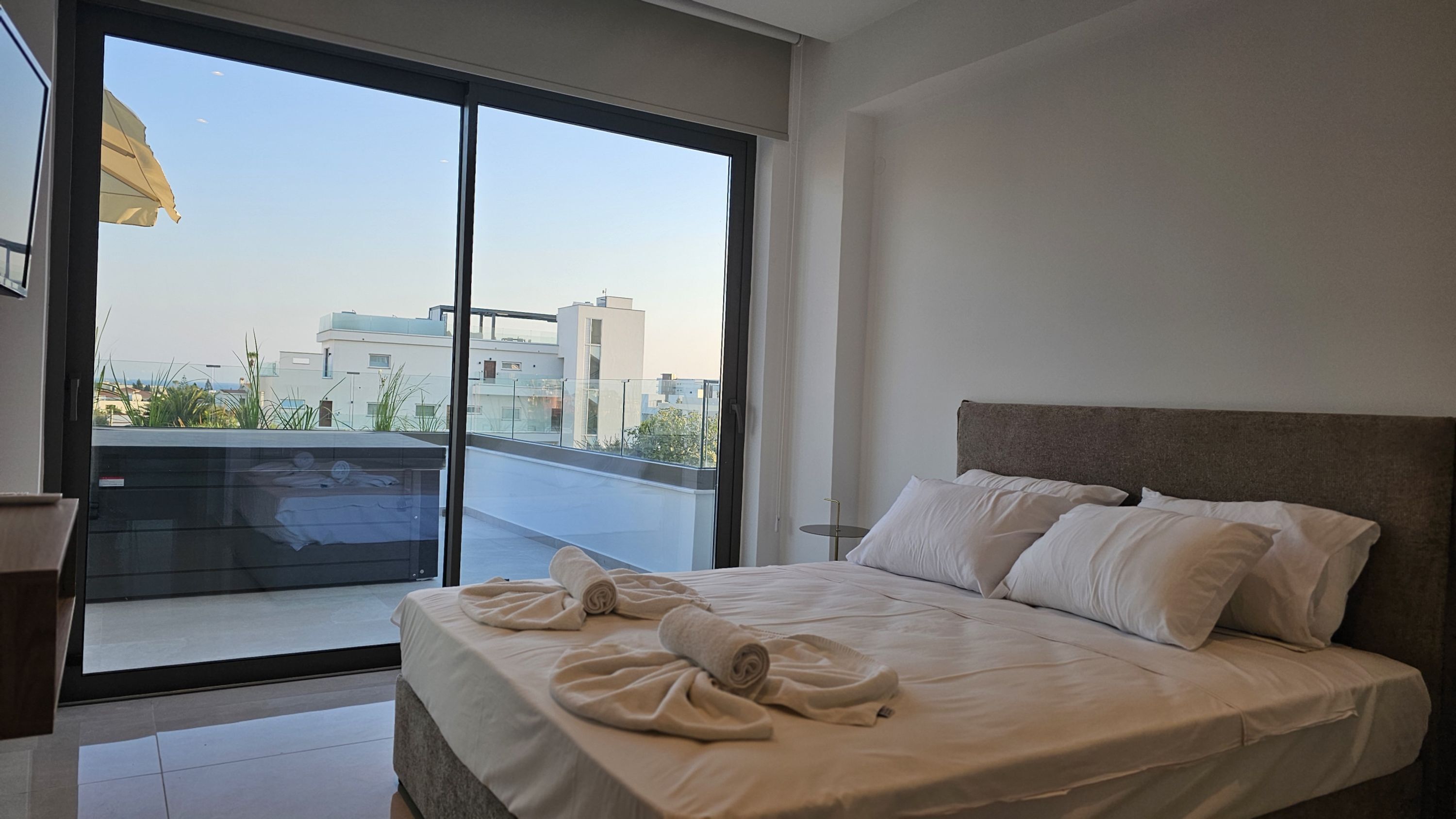 Une chambre d’hôtel moderne dotée d’un grand lit, offrant une vue sur la ville grâce à de larges fenêtres du sol au plafond, et d’un balcon avec une balustrade en verre.