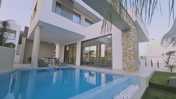 Une maison moderne dotée d’une piscine, d’un patio avec des chaises et d’un pilier en pierre.